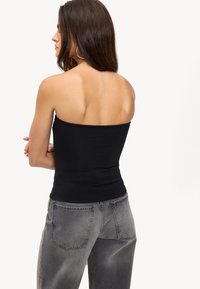 Femme aux longs cheveux noirs, portant un top noir sans bretelles et un jean gris délavé, debout les bras croisés, dos à l'appareil photo.