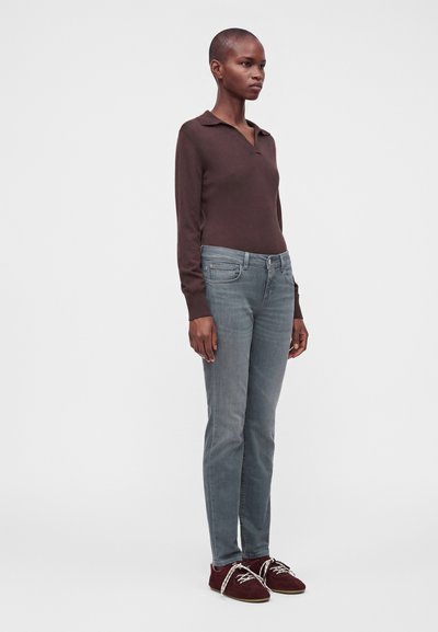 Personne debout portant un haut marron à manches longues, un jean gris clair et des chaussures à lacets rouge foncé sur un fond clair uni.