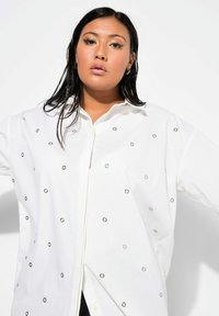 Camisa blanca de botones con acentuaciones en grommets plateados, corte holgado y cuello clásico. Tejido suave y diseño estructurado.