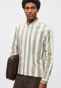 Πουκάμισο με μακριά μανίκια, με κάθετες ρίγες σε πράσινο ελιάς και λευκό, γιακά με κουμπιά, μπροστινή τσέπη, φορεμένο με καφέ τσάντα ταχυδρόμου.