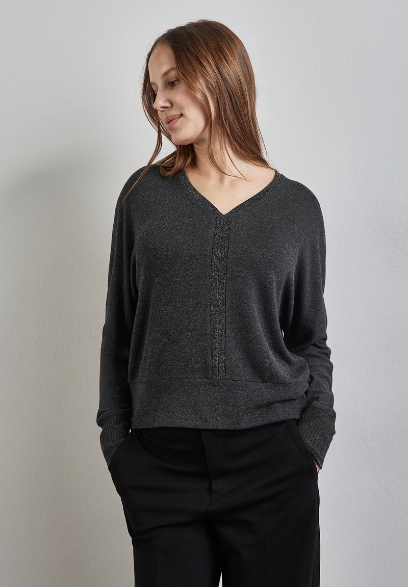 Pull gris à col en V avec des poignets et un ourlet côtelés, présentant un subtil détail de couture verticale et une coupe décontractée. Assorti à un pantalon noir.