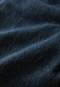 Tejido de denim oscuro y texturizado con sutiles motas, presenta una superficie lisa y un brillo ligeramente reflectante. Pliegues curvos crean profundidad.