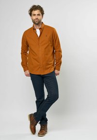 Roodbruine button-up overhemd, van katoen, lange mouwen, gecombineerd met donkere spijkerbroek en bruine veterschoenen. Eenvoudig logo-detail op het overhemd.