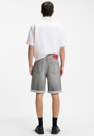 Homme portant une chemise blanche à manches courtes et un short en jean gris avec un ourlet roulé et une étiquette rouge "Hugo", chaussé de chaussettes noires et de chaussures noires, debout de dos.
