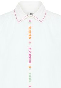 Chemise blanche à col avec surpiqûres roses sur le col et broderie florale verticale formant le mot « FLOWERS » en rose, orange et vert le long de la ligne de boutons.