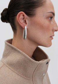 Metallisch zilveren oorbellen met een langwerpige, druppelvorm, gladde textuur. Gedragen met een beige, hoog gesloten, wolachtige jas met drukknopen.