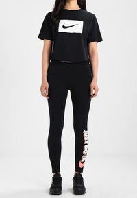 Frau in schwarzem Nike-Cropped-T-Shirt und schwarzen Leggings mit pinkem "Just Do It"-Text sowie schwarzen Sneakers vor weißem Hintergrund.