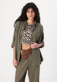 Camisa verde oliva com botões sobre um top cropped de estampa de leopardo, combinada com calças de perna larga e um cinto castanho com detalhes em metal em forma circular.