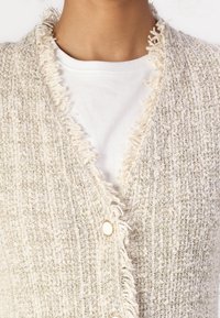 Cardigan texturé beige avec des bords effilochés, featuring un col en V et une fermeture à un bouton sur un t-shirt blanc.