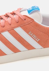 Sneaker in suede rosa con accenti in pelle bianca e tre strisce bianche. Presenta un logo sul lato e una suola in gomma testurizzata.