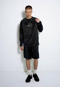 Schwarzes Sportoutfit mit einem Langarmshirt aus strukturiertem Stoff und Shorts. Enthält ein auffälliges Logo und schwarze Schuhe mit dicken Sohlen.