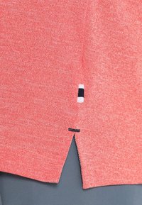 Korallinpunainen neulottu kangas, jossa pieni sivuhalkio ja valkoinen sekä laivastonsininen suorakulmainen lappu helman lähellä tummanharmaiden housujen päällä.