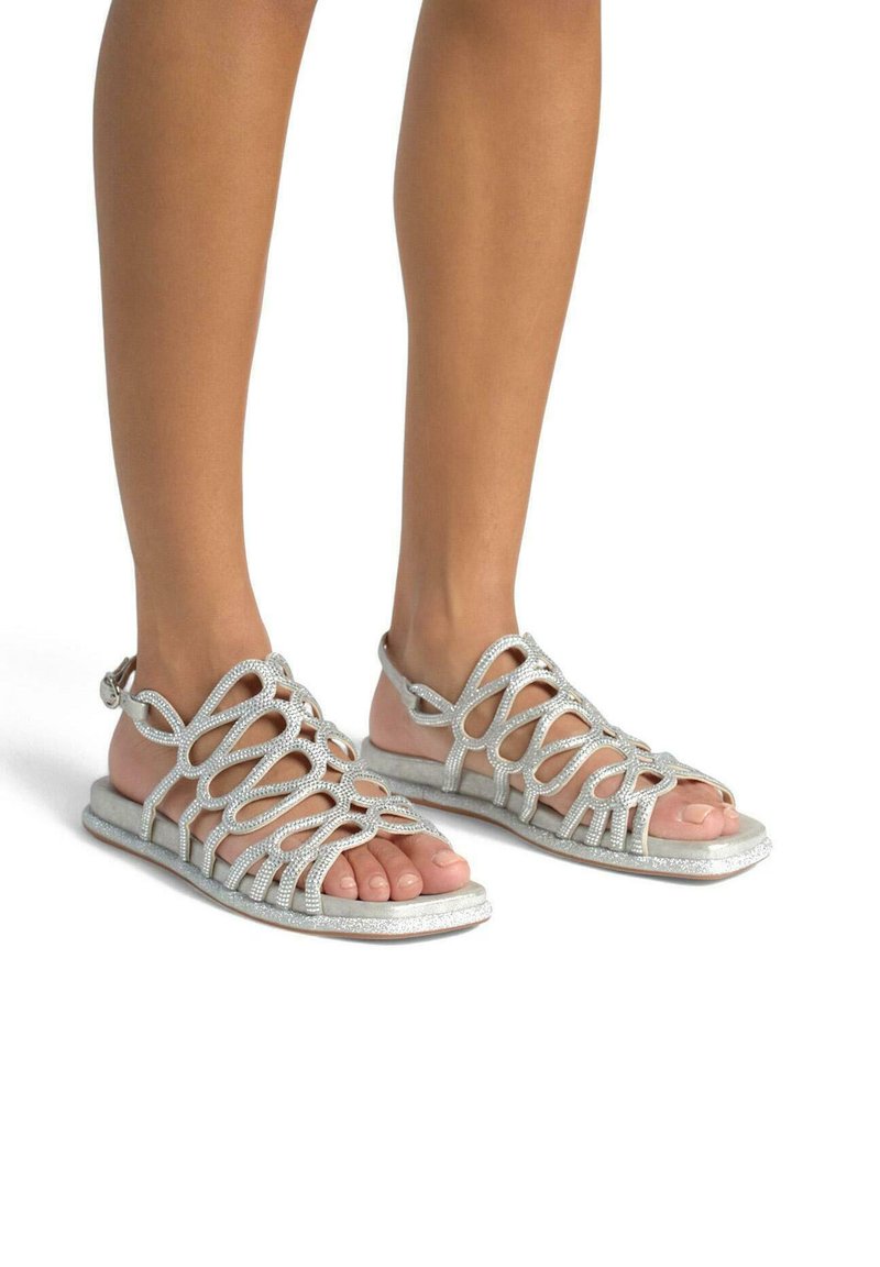 Srebrne, błyszczące płaskie sandały z krzyżowym wzorem i odkrytymi palcami. Posiadają pasek na kostkę i teksturowaną podeszwę.