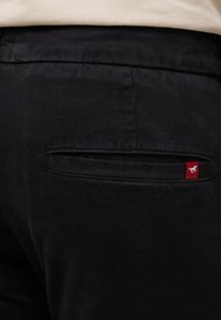 Czarne spodnie z tylną kieszenią z czerwoną metką. Materiał wydaje się gładki z lekkim połyskiem, co podkreśla minimalistyczny design.