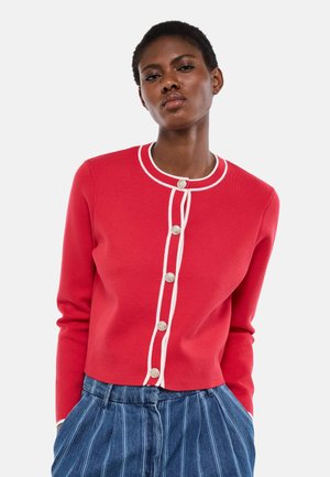 Femme portant un cardigan rouge à boutons avec des bordures blanches et un pantalon taille haute bleu à rayures verticales, debout devant un fond uni.