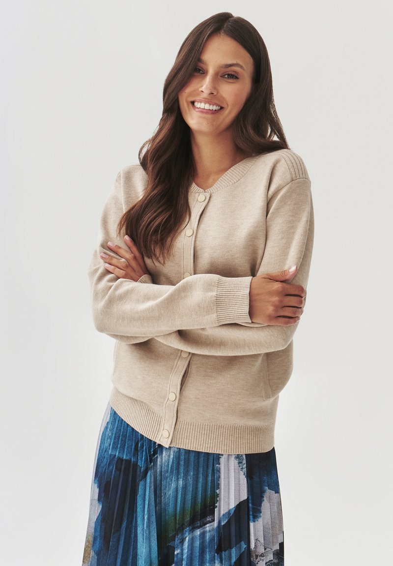 Beige cardigan met een ronde halslijn, geribbelde accenten en knoopsluiting. Gecombineerd met een geplooide, kleurrijk patroonrok.