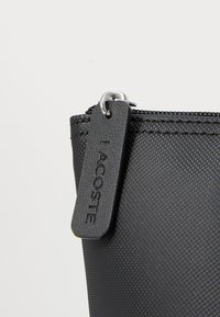 Punga neagră cu fermoar, realizată dintr-un material texturat, cu o etichetă atârnată pe care scrie "LACOSTE". Are un design simplu și funcțional.