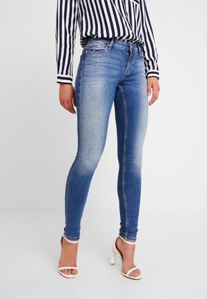 Frau trägt blaue ausgewaschene Skinny-Jeans, ein schwarz-weiß gestreiftes Langarmshirt und weiße Sandalen mit hohem Absatz, steht auf weißem Boden.