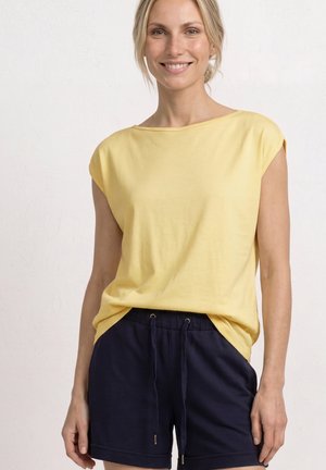 Femme souriante portant un haut jaune sans manches ample et un short bleu marine à cordon, debout devant un fond clair uni.
