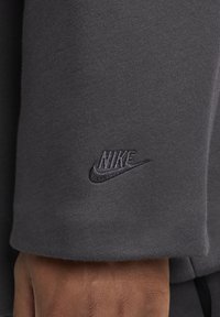 Szara rękaw bluzy z teksturowanym wykończeniem; na górnej części ramienia znajduje się ciemne, haftowane logo Nike. Gładki bawełniany materiał.