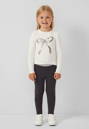 Camicia bianca a maniche lunghe con una grafica di nastri argentati al centro, abbinata a leggins grigio scuro e sneaker bianche. Tessuto dalla consistenza liscia.
