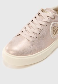 Svetloružičasta metalik tenisica s odgovarajućim vezicama, bijelim potplatom i ukrasnim grbom sa strane.