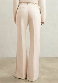 Osoba v béžových nohaviciach s vysokým pásom a širokými nohavicami a svetlom béžovom svetri, stojaca na hladkej betónovej podlahe pred obyčajnou stenou.