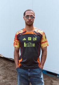 Grå sportsjakke med røde detaljer, skarpe orange og gule mønstrede ærmer, sorte grafikker og logoer. Båret sammen med blå jeans.