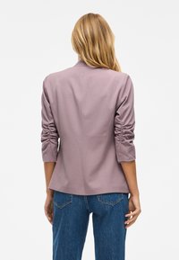 Blazer de lavanda con un diseño entallado, mangas fruncidas y tejido suave. Combinado con jeans azules de cintura alta.