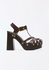 Sandale compensée en cuir marron avec talon haut épais, sangle de cheville à boucle dorée et accents de rivets dorés sur les sangles et la semelle.