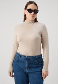 Bežna dolga majica s turtleneck ovratnikom iz mehkega materiala, v kombinaciji z modrimi hlačami z visokim pasom, ki imajo zapiranje na gumb in sprednja žepa.