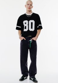 Černý oversized dres s bílým číslem "80" a pruhovanými rukávy, kombinovaný s tmavě fialovými džíny a černými botami s texturovaným povrchem.