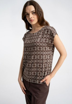 Kobieta w brązowej bluzce z krótkim rękawem z białym geometrycznym wzorem i ciemnobrązowych spodniach, stojąca jedną ręką w kieszeni na jednolitym tle.