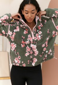 Fleece mikina olivové barvy s růžovým květinovým vzorem, vpředu na půl zip, s růžovými detaily; má měkký, texturovaný povrch.