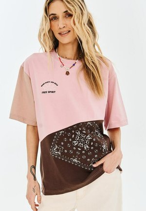 Kobieta o falowanych blond włosach nosi oversize'owy, różowo-brązowy T-shirt w kolorowe bloki z napisem "Free Spirit" i kwiatowym wzorem, beżowe spodnie oraz naszyjnik.