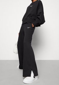 Czarna bluza Nike z okrągłym dekoltem, luźny krój. Pasujące czarne spodnie dresowe z bocznymi kieszeniami, rozcięty brzeg i białe logo na obu. Białe trampki z czarnymi akcentami.