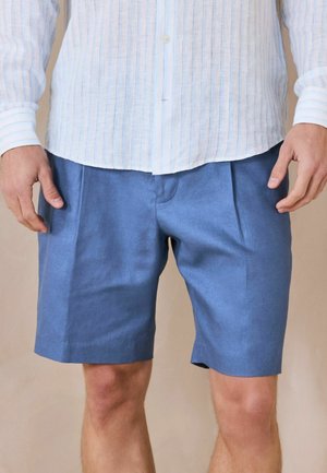 Bărbat purtând o cămașă cu mâneci lungi, cu dungi albastre deschise, și pantaloni scurți croiți, albaștri, stând cu mâinile relaxate pe lângă corp, pe un fundal neutru.