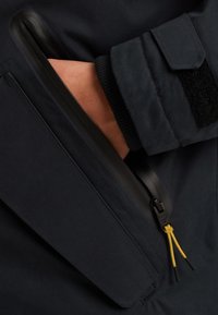 Námorní plášť s kapsou na zip, se černým zipem a žlutými táhly. Materiál vypadá hladce s jemnými texturovými detaily.