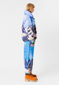 Chaqueta acolchada en tonos azules con estampado de montañas y texto blanco, combinada con pantalones texturizados azules a juego y botas naranjas con suelas gruesas.