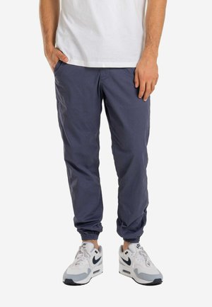 Mann trägt marineblaue Jogginghosen, weiße Sneakers mit schwarzem Nike-Swoosh und ein weißes T-Shirt, Hände lässig in den Taschen.