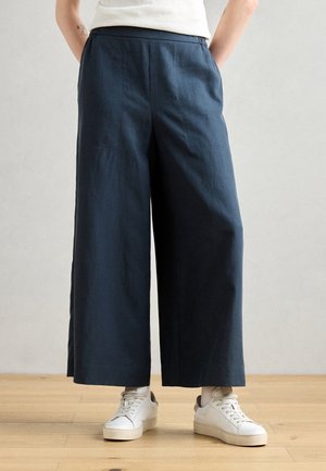 Person trägt marineblaue, weit geschnittene, verkürzte Hosen und weiße Turnschuhe, steht auf einem Holzboden vor einer schlichten grauen Wand.