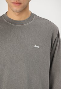 Grå langermet skjorte med rund hals og ribbestrikket krage, med en liten hvit "obey"-logo på brystet. Mykt bomullsmateriale.