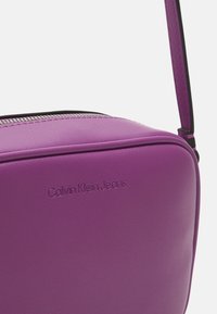 Borsa a tracolla in pelle viola con logo Calvin Klein Jeans in rilievo e zip argentata visibile vicino al bordo superiore.
