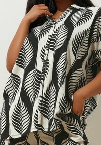 Blusa bianca e nera con stampa a foglie, maniche corte, chiusura con bottoni e tessuto strutturato. Le tasche ai lati aumentano la funzionalità.