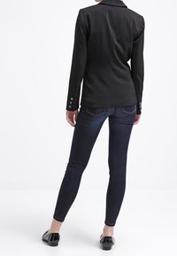 Blazer noir aux épaules structurées, fermeture à un bouton et tissu texturé, associé à un jean skinny bleu foncé et des ballerines noires.