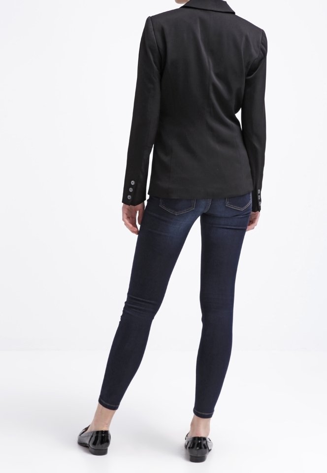 Blazer noir aux épaules structurées, fermeture à un bouton et tissu texturé, associé à un jean skinny bleu foncé et des ballerines noires.