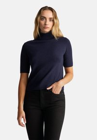 Mladá žena s blond vlasy, oblečená v námořnickém svetru s krátkým rukávem a černých kalhotách, stojící s jednou rukou v kapse.