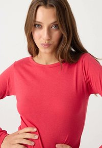 Camiseta de manga larga con cuello redondo en tejido rojo vibrante. Textura suave con detalles de costuras visibles. Modelo posando con las manos en las caderas.