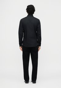 Άνδρας με σγουρά μαλλιά φορώντας μαύρο πουκάμισο μακρυμάνικο και μαύρο παντελόνι, στέκεται γυρισμένος με την πλάτη σε ένα απλό ανοιχτό φόντο.