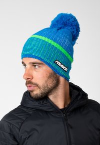 Blue knit beanie with a green stripe, pom-pom on top, and textured pattern. Features a black branded patch. Worn by a man in a black jacket.